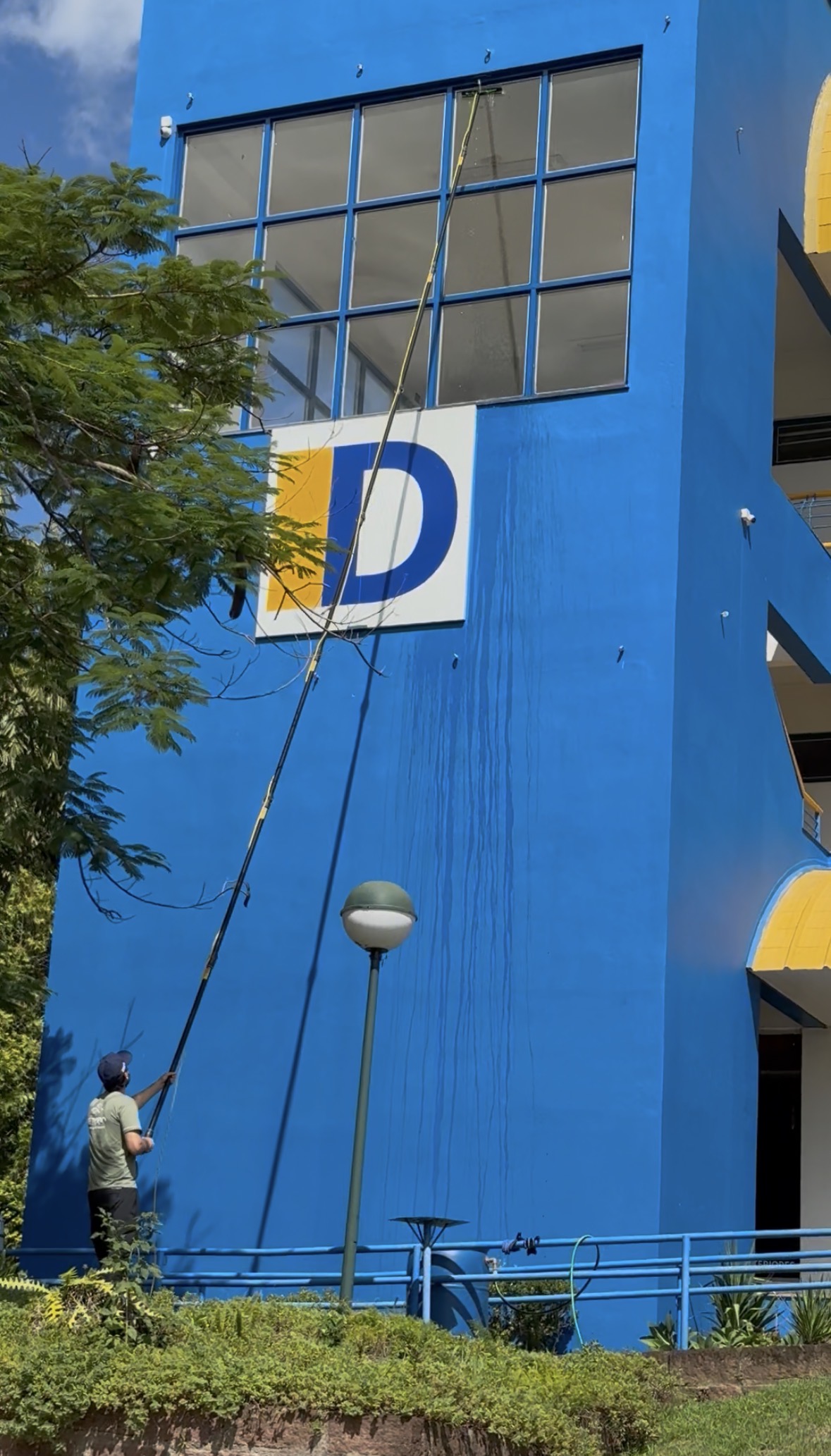Técnico realizando limpeza de vidros em fachada azul com equipamento de alcance.